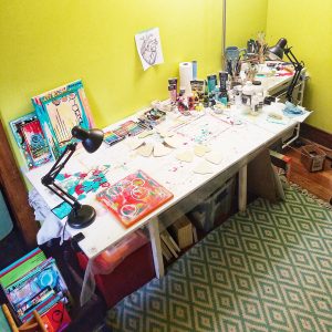 Rebecca Weger's home painting sudio. Her painting table, with a couple of in-process paintings, and a collection o fpaints, inks, mediums and brushes.